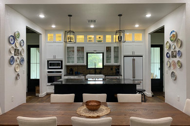 Example of a cottage medium tone wood floor and brown floor kitchen design in Houston with a farmhouse sink, white cabinets, granite countertops, metallic backsplash, metal backsplash, white appliances, an island and black countertops