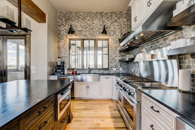 Mid-sized urban l-shaped light wood floor and brown floor open concept kitchen photo in Dallas with gray backsplash, stainless steel appliances, an island, a farmhouse sink, recessed-panel cabinets, white cabinets, granite countertops, subway tile backsplash and black countertops