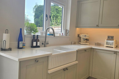 Photo of a medium sized contemporary l-shaped kitchen/diner in West Midlands with a belfast sink, shaker cabinets, beige cabinets, quartz worktops, white splashback, stone slab splashback, black appliances, medium hardwood flooring, no island, grey floors, white worktops and a coffered ceiling.