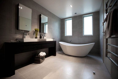 This is an example of a large contemporary master bathroom in Auckland with a vessel sink, furniture-like cabinets, dark wood cabinets, wood benchtops, a freestanding tub, a curbless shower, a one-piece toilet, porcelain tile, porcelain floors, gray tile and grey floor.