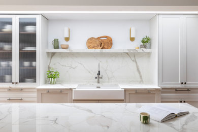 Photo of a large traditional galley kitchen/diner in London with a belfast sink, shaker cabinets, white cabinets, composite countertops, white splashback, porcelain splashback, stainless steel appliances, medium hardwood flooring, an island, white worktops and feature lighting.