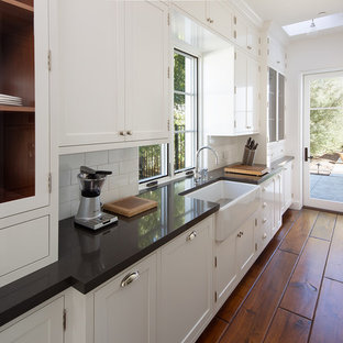 Inspiration for a mediterranean galley kitchen pantry in San Diego with a farmhouse sink, shaker cabinets, white cabinets, white splashback, subway tile splashback, medium hardwood floors, no island, brown floor and black benchtop.