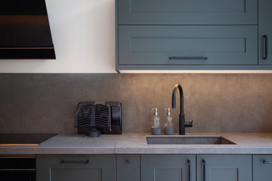 This is an example of a large modern grey and black l-shaped open plan kitchen in Oxfordshire with a single-bowl sink, shaker cabinets, green cabinets, quartz worktops, grey splashback, porcelain splashback, black appliances, porcelain flooring, an island, beige floors, white worktops, exposed beams and a chimney breast.