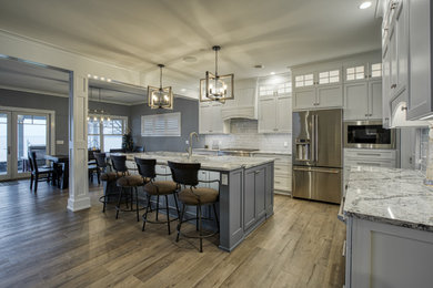 This is an example of an arts and crafts eat-in kitchen in Other with a farmhouse sink, shaker cabinets, white cabinets, quartzite benchtops, grey splashback, stainless steel appliances, medium hardwood floors, with island, brown floor and multi-coloured benchtop.