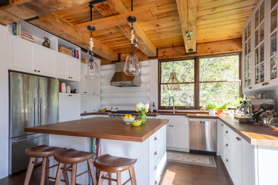Example of a mid-sized classic u-shaped medium tone wood floor, brown floor and exposed beam eat-in kitchen design in Montreal with a farmhouse sink, shaker cabinets, white cabinets, wood countertops, white backsplash, wood backsplash, stainless steel appliances, an island and brown countertops