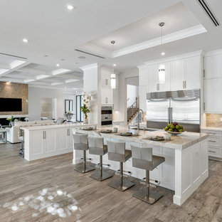 Photo of a large transitional l-shaped open plan kitchen in Miami with a farmhouse sink, beaded inset cabinets, white cabinets, quartzite benchtops, beige splashback, granite splashback, stainless steel appliances, light hardwood floors, multiple islands, beige floor, white benchtop and coffered.