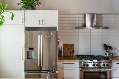 Large urban single-wall light wood floor open concept kitchen photo in Philadelphia with a drop-in sink, flat-panel cabinets, white cabinets, wood countertops, white backsplash, stainless steel appliances, an island and ceramic backsplash