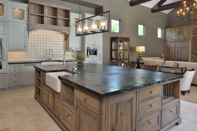 Design ideas for a large traditional u-shaped open plan kitchen in Houston with with island, raised-panel cabinets, medium wood cabinets, soapstone benchtops, beige splashback, ceramic splashback, stainless steel appliances, a farmhouse sink and travertine floors.