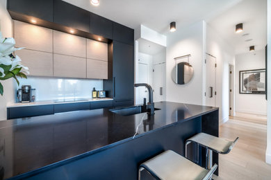 Example of a trendy galley laminate floor and beige floor eat-in kitchen design in Vancouver with an undermount sink, flat-panel cabinets, black cabinets, quartz countertops, white backsplash, quartz backsplash, black appliances, an island and black countertops