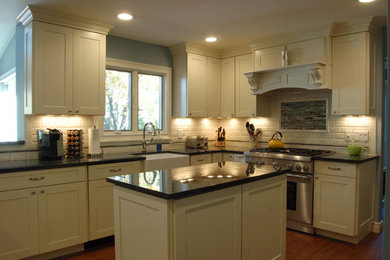Example of a classic u-shaped eat-in kitchen design in New York with a farmhouse sink, shaker cabinets, yellow cabinets, granite countertops, white backsplash, porcelain backsplash and stainless steel appliances