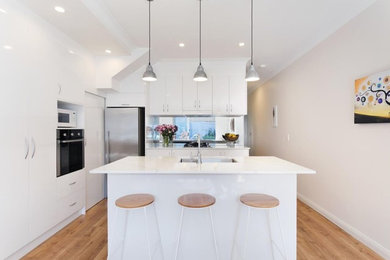 Photo of a mid-sized modern galley open plan kitchen in Sydney with an undermount sink, beaded inset cabinets, white cabinets, glass sheet splashback, panelled appliances, light hardwood floors and multiple islands.