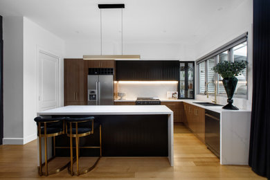 This is an example of a large contemporary eat-in kitchen in Melbourne with a double-bowl sink, flat-panel cabinets, dark wood cabinets, quartz benchtops, white splashback, engineered quartz splashback, stainless steel appliances, light hardwood floors, with island and white benchtop.