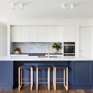 Inspiration for a mid-sized beach style open plan kitchen in Sydney with a drop-in sink, shaker cabinets, white cabinets, marble benchtops, grey splashback, marble splashback, black appliances, medium hardwood floors, with island, brown floor, grey benchtop and timber.