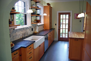 This is an example of a mid-sized arts and crafts galley kitchen in Other with a farmhouse sink, shaker cabinets, light wood cabinets, soapstone benchtops, beige splashback, stone tile splashback, stainless steel appliances, linoleum floors, with island and black floor.