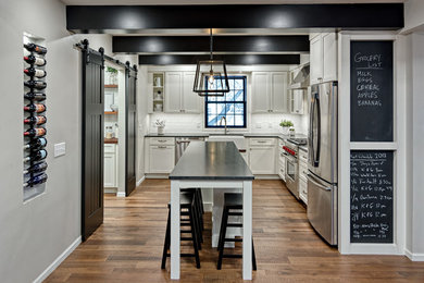 Photo of a transitional l-shaped kitchen in Other with a farmhouse sink, shaker cabinets, white cabinets, white splashback, subway tile splashback, stainless steel appliances, medium hardwood floors, with island, brown floor and black benchtop.