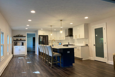 Example of a large transitional l-shaped vinyl floor and brown floor kitchen pantry design in Cleveland with a farmhouse sink, shaker cabinets, white cabinets, quartz countertops, white backsplash, subway tile backsplash, black appliances, an island and white countertops