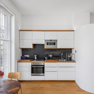 Новый формат декора квартиры: маленькая линейная кухня в современном стиле с обеденным столом, накладной раковиной, плоскими фасадами, белыми фасадами, столешницей из дерева, фартуком из сланца, черным фартуком, техникой из нержавеющей стали и паркетным полом среднего тона