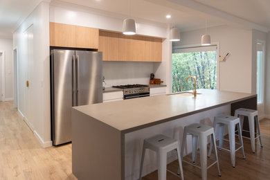 Large modern galley open plan kitchen in Melbourne with an undermount sink, flat-panel cabinets, light wood cabinets, solid surface benchtops, white splashback, glass sheet splashback, stainless steel appliances, light hardwood floors, with island, beige floor and grey benchtop.