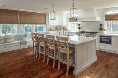 Huge elegant l-shaped medium tone wood floor and brown floor eat-in kitchen photo in New York with a farmhouse sink, shaker cabinets, white cabinets, solid surface countertops, white backsplash, ceramic backsplash, stainless steel appliances, an island and white countertops
