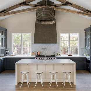 Diseño de cocina en U, clásica renovada, grande, abierta, con fregadero bajoencimera, armarios estilo shaker, puertas de armario azules, salpicadero de losas de piedra, electrodomésticos de acero inoxidable, suelo de madera clara, una isla, encimera de mármol, salpicadero blanco y suelo marrón