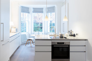 Design ideas for a medium sized open plan kitchen in London with flat-panel cabinets, white cabinets, marble worktops, white splashback, stainless steel appliances, medium hardwood flooring, an island, brown floors, white worktops and feature lighting.