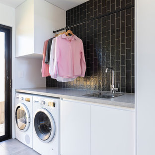 Photo of a medium sized contemporary single-wall utility room in Brisbane with a single-bowl sink, flat-panel cabinets, white cabinets, engineered stone countertops, black walls, ceramic flooring, a side by side washer and dryer, grey floors and grey worktops.