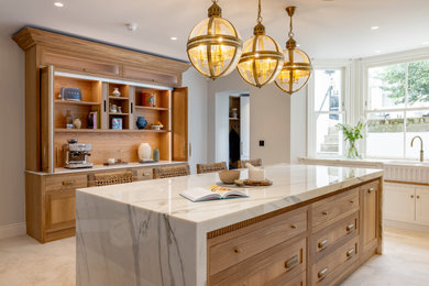 Photo of a large traditional open plan kitchen in London with a belfast sink, shaker cabinets, light wood cabinets, marble worktops, white splashback, marble splashback, porcelain flooring, an island, beige floors and white worktops.