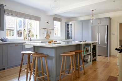 Large l-shaped separate kitchen in West Midlands with a farmhouse sink, shaker cabinets, grey cabinets, solid surface benchtops, stainless steel appliances, light hardwood floors, with island and white benchtop.