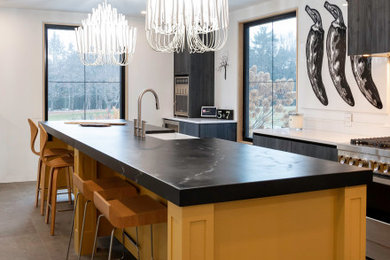 Eat-in kitchen - large transitional galley ceramic tile and gray floor eat-in kitchen idea in Toronto with a farmhouse sink, flat-panel cabinets, yellow cabinets, soapstone countertops, white backsplash, porcelain backsplash, paneled appliances, an island and black countertops