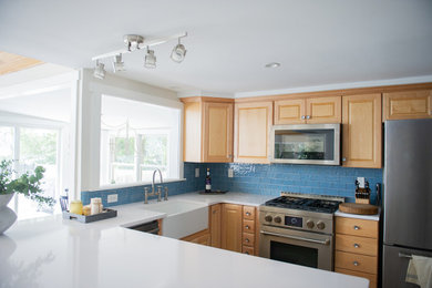 Inspiration for a mid-sized coastal l-shaped bamboo floor and beige floor enclosed kitchen remodel in New York with a farmhouse sink, raised-panel cabinets, light wood cabinets, solid surface countertops, blue backsplash, ceramic backsplash, stainless steel appliances, a peninsula and white countertops