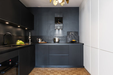This is an example of a scandinavian l-shaped kitchen in Paris with a single-bowl sink, flat-panel cabinets, black cabinets, black splashback, black appliances, light hardwood floors and black benchtop.