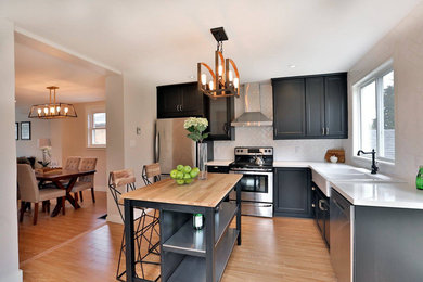 Open concept kitchen - mid-sized transitional l-shaped light wood floor open concept kitchen idea in Toronto with a farmhouse sink, shaker cabinets, black cabinets, quartz countertops, white backsplash, subway tile backsplash, stainless steel appliances and an island