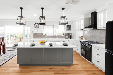 Photo of a large traditional l-shaped eat-in kitchen in Perth with shaker cabinets, white cabinets, brown splashback, black appliances, medium hardwood floors, multiple islands, brown floor, white benchtop, a farmhouse sink, quartz benchtops and ceramic splashback.