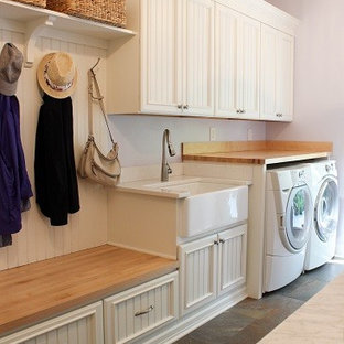 Photo of a medium sized traditional single-wall utility room in Other with a belfast sink, recessed-panel cabinets, white cabinets, wood worktops, purple walls, porcelain flooring, a side by side washer and dryer and grey floors.
