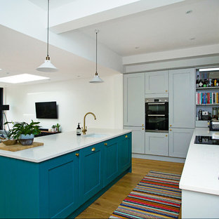 Photo of a large contemporary l-shaped kitchen/diner in London with a submerged sink, shaker cabinets, grey cabinets, quartz worktops, green splashback, ceramic splashback, an island, white worktops, black appliances, medium hardwood flooring and brown floors.