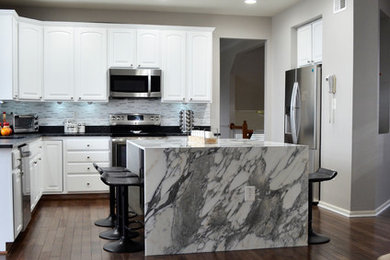Example of a large trendy l-shaped medium tone wood floor and brown floor open concept kitchen design in DC Metro with a double-bowl sink, raised-panel cabinets, white cabinets, marble countertops, yellow backsplash, stone tile backsplash, stainless steel appliances and an island