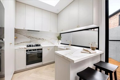 Inspiration for a small modern l-shaped light wood floor and beige floor eat-in kitchen remodel in Los Angeles with a farmhouse sink, shaker cabinets, white cabinets, marble countertops, beige backsplash, marble backsplash, white appliances, a peninsula and beige countertops