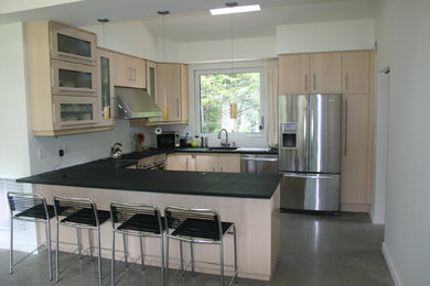 Example of a small trendy u-shaped concrete floor kitchen design in New York with an undermount sink, flat-panel cabinets, light wood cabinets, soapstone countertops, white backsplash, a peninsula and stainless steel appliances