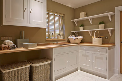 Photo of a country l-shaped utility room in Wiltshire with an undermount sink, shaker cabinets, white cabinets, wood benchtops, beige walls, grey floor and beige benchtop.