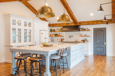 Inspiration for a large transitional l-shaped eat-in kitchen in Detroit with glass-front cabinets, white cabinets, with island, a farmhouse sink, white appliances, white benchtop, light hardwood floors, brown floor, exposed beam, white splashback and subway tile splashback.