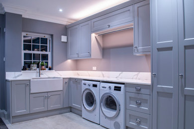 Photo of a large traditional l-shaped utility room in Essex with a belfast sink, flat-panel cabinets, grey cabinets, composite countertops, grey walls, travertine flooring, a side by side washer and dryer, beige floors, white worktops and feature lighting.