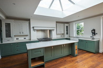 Inspiration for a large classic open plan kitchen in London with a belfast sink, shaker cabinets, green cabinets, quartz worktops, metallic splashback, stainless steel appliances, dark hardwood flooring and an island.