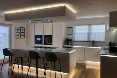 Large contemporary l-shaped eat-in kitchen in Glasgow with an integrated sink, flat-panel cabinets, white cabinets, quartzite benchtops, green splashback, glass sheet splashback, black appliances, vinyl floors, with island, brown floor, white benchtop and recessed.