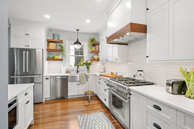 Mid-sized transitional l-shaped kitchen in Providence with a farmhouse sink, shaker cabinets, white cabinets, quartz benchtops, white splashback, ceramic splashback, stainless steel appliances, medium hardwood floors, a peninsula, brown floor and white benchtop.