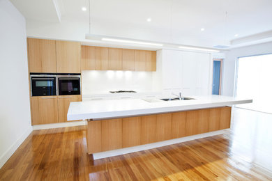 Large contemporary galley open plan kitchen in Adelaide with a drop-in sink, flat-panel cabinets, light wood cabinets, quartz benchtops, white splashback, glass sheet splashback, black appliances, medium hardwood floors and with island.