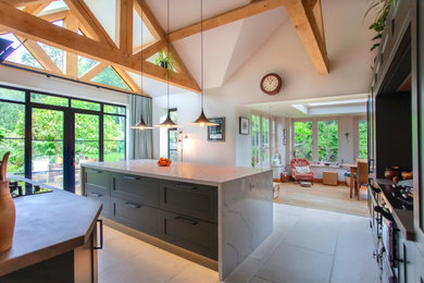 Large modern grey and black l-shaped open plan kitchen in Oxfordshire with a single-bowl sink, shaker cabinets, green cabinets, quartz worktops, grey splashback, porcelain splashback, black appliances, porcelain flooring, an island, beige floors, white worktops, exposed beams and a chimney breast.