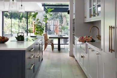Inspiration for a large single-wall eat-in kitchen in London with a farmhouse sink, raised-panel cabinets, white cabinets, solid surface benchtops, white splashback, ceramic splashback, stainless steel appliances, light hardwood floors, with island, brown floor and white benchtop.