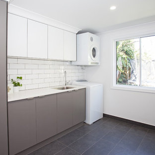 Ejemplo de cuarto de lavado lineal, exótico, grande, con fregadero bajoencimera, armarios con paneles lisos, puertas de armario grises, encimera de cuarzo compacto, paredes blancas, lavadora y secadora apiladas y suelo negro