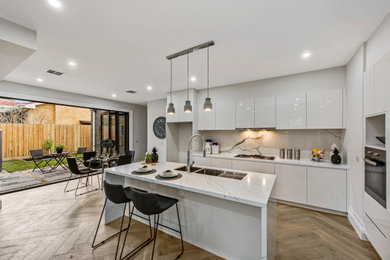 Mid-sized contemporary u-shaped eat-in kitchen in Melbourne with a drop-in sink, recessed-panel cabinets, white cabinets, quartzite benchtops, multi-coloured splashback, stone slab splashback, stainless steel appliances, medium hardwood floors, with island, beige floor and white benchtop.