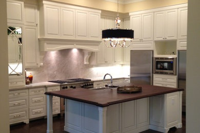Large trendy l-shaped dark wood floor open concept kitchen photo in Houston with a farmhouse sink, raised-panel cabinets, white cabinets, wood countertops, gray backsplash, stone slab backsplash, stainless steel appliances and an island